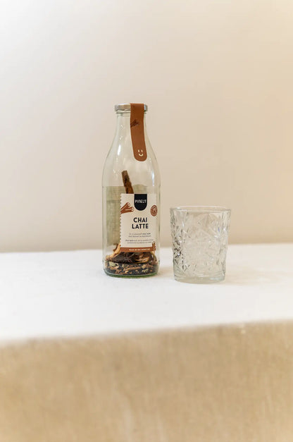 De glazen fles van Pineut is voor jou alvast gevuld met alle kruiden en specerijen die je nodig hebt om zelf een heerlijke Chai Latte te maken. Dit sfeerbeeld toont de fraaie flex op een naturel tafelkleed met een mooi glas ernaast.